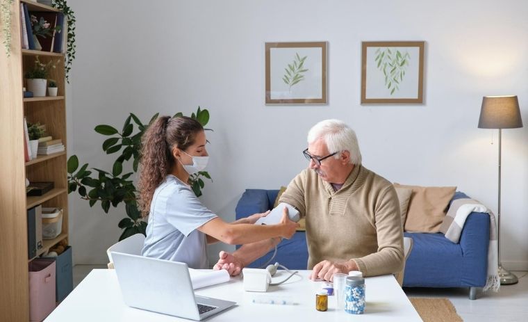 A nurse from a Home Health Care Agency in New York checking an elderly man’s blood pressure at home during a caregiving visit.