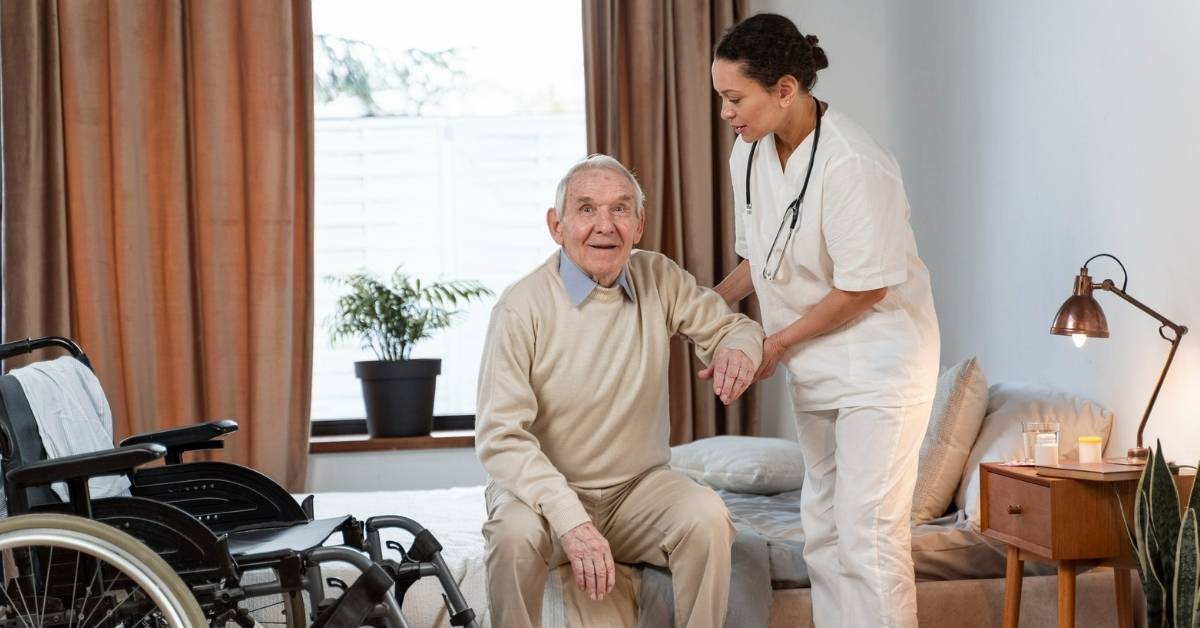 Registered nurse providing skilled nursing care at home, assisting an elderly man to stand beside his wheelchair in a bright New York apartment.